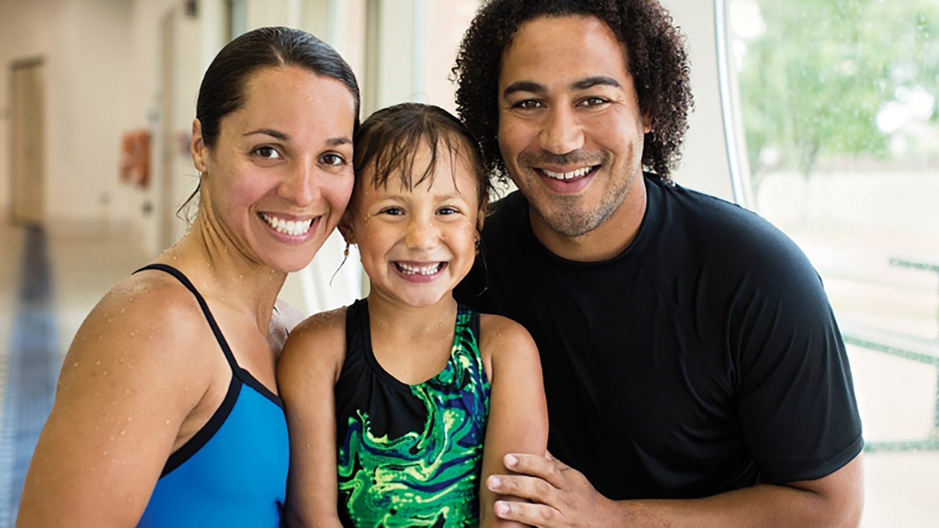 Smiling mother, young daughter, and father together at an indoor YMCA pool wearing swimsuits