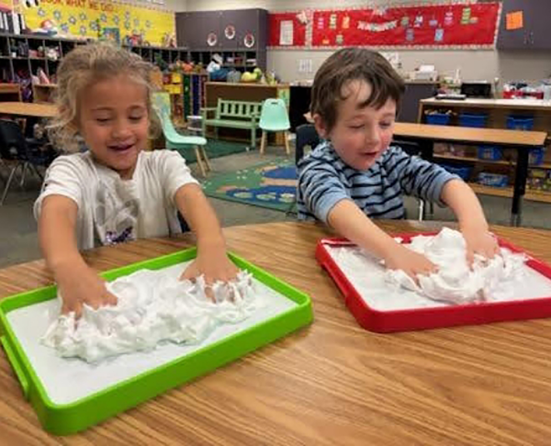 Two young children playing with shaving cream on colorful trays in a preschool classroom