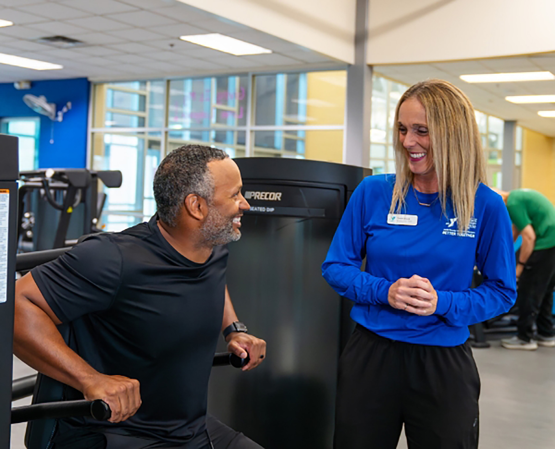 Smiling YMCA trainer in a blue staff shirt coaching a man using a strength machine in the gym