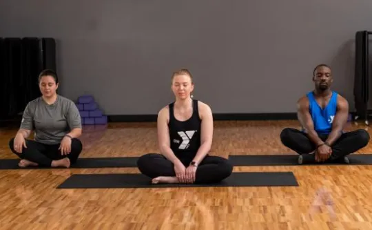 Three adults seated cross-legged on yoga mats meditating in a studio, center person wearing a YMCA tank top