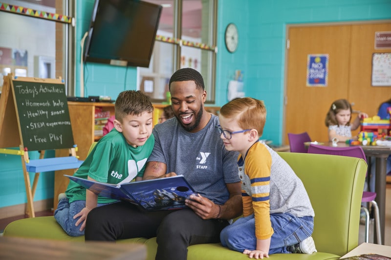 YMCA staff member reading a book with two young boys on a green couch