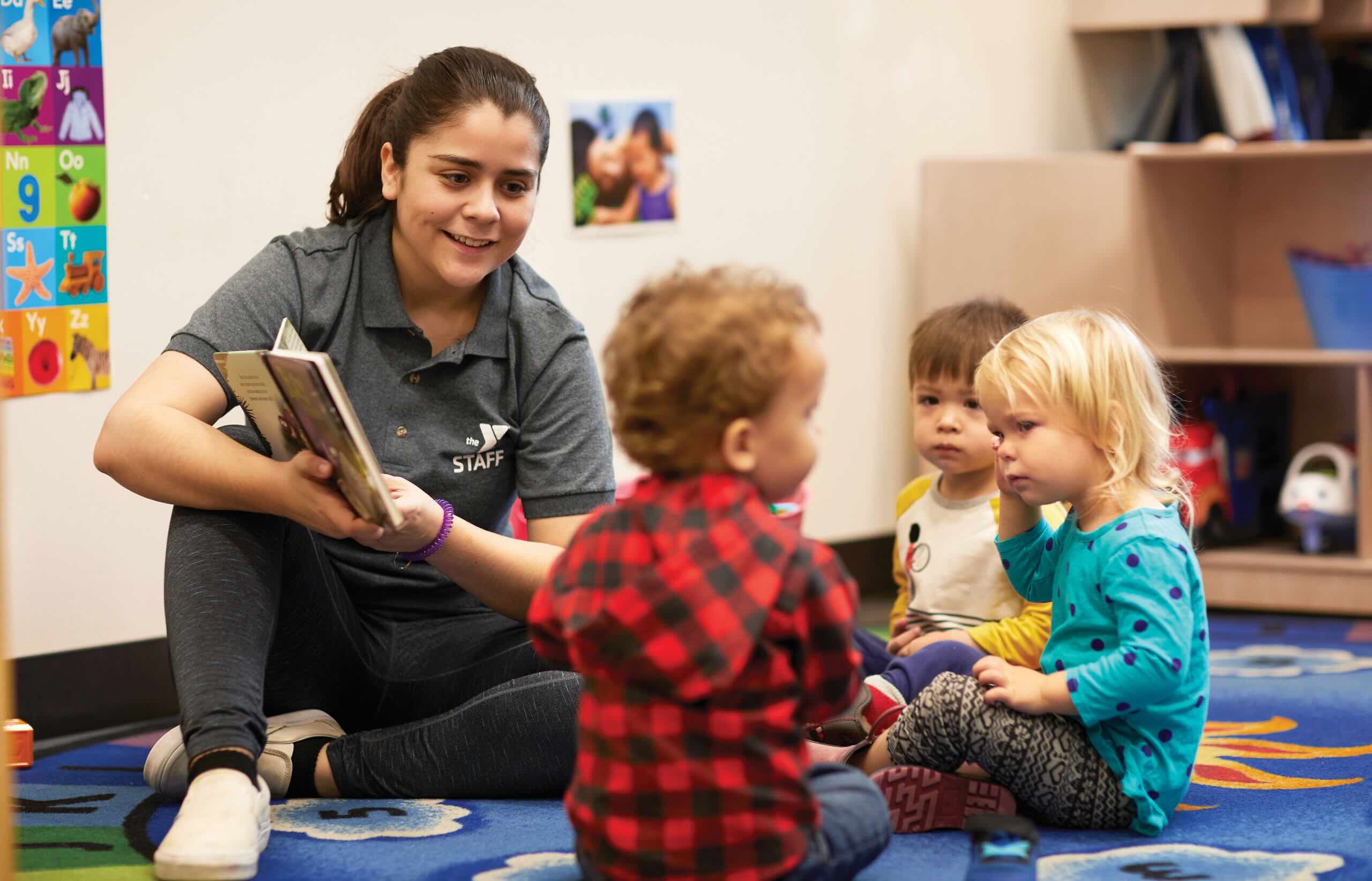 YMCA staff member in a staff polo reads a picture book to toddlers on a classroom rug