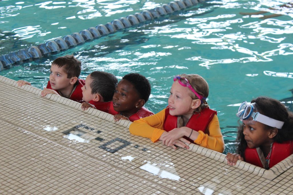 Five children in red life jackets and goggles line the edge of an indoor pool