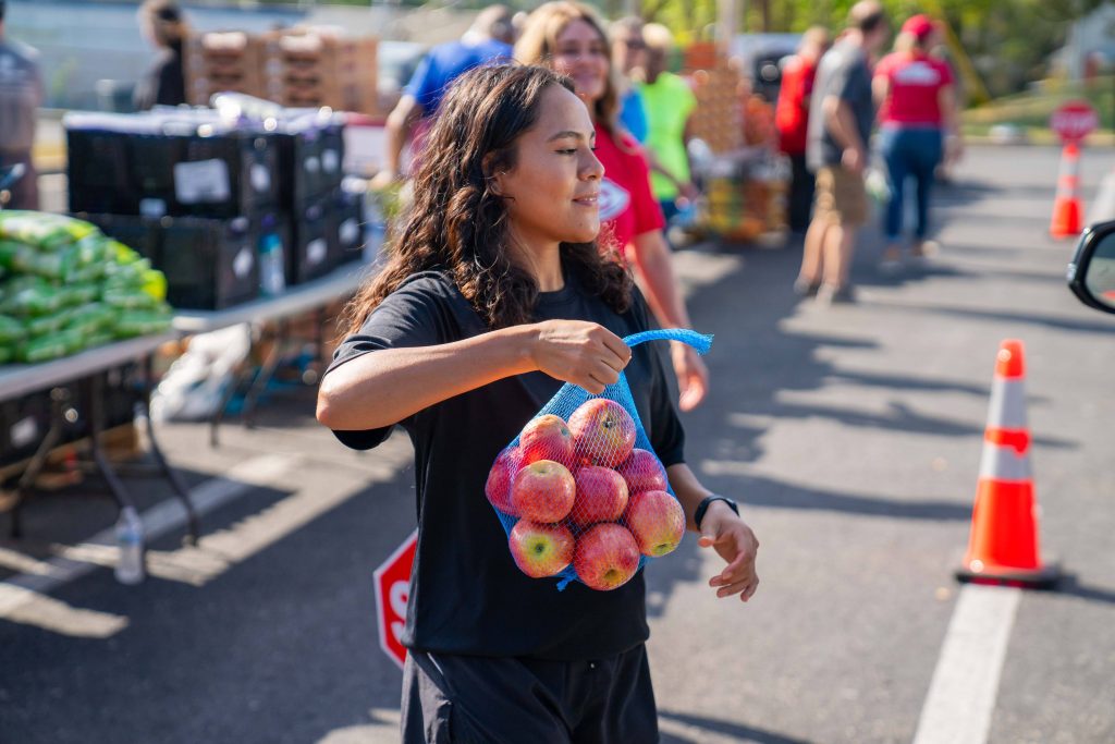 Smiling volunteer carrying a mesh bag of apples at an outdoor community food distribution event