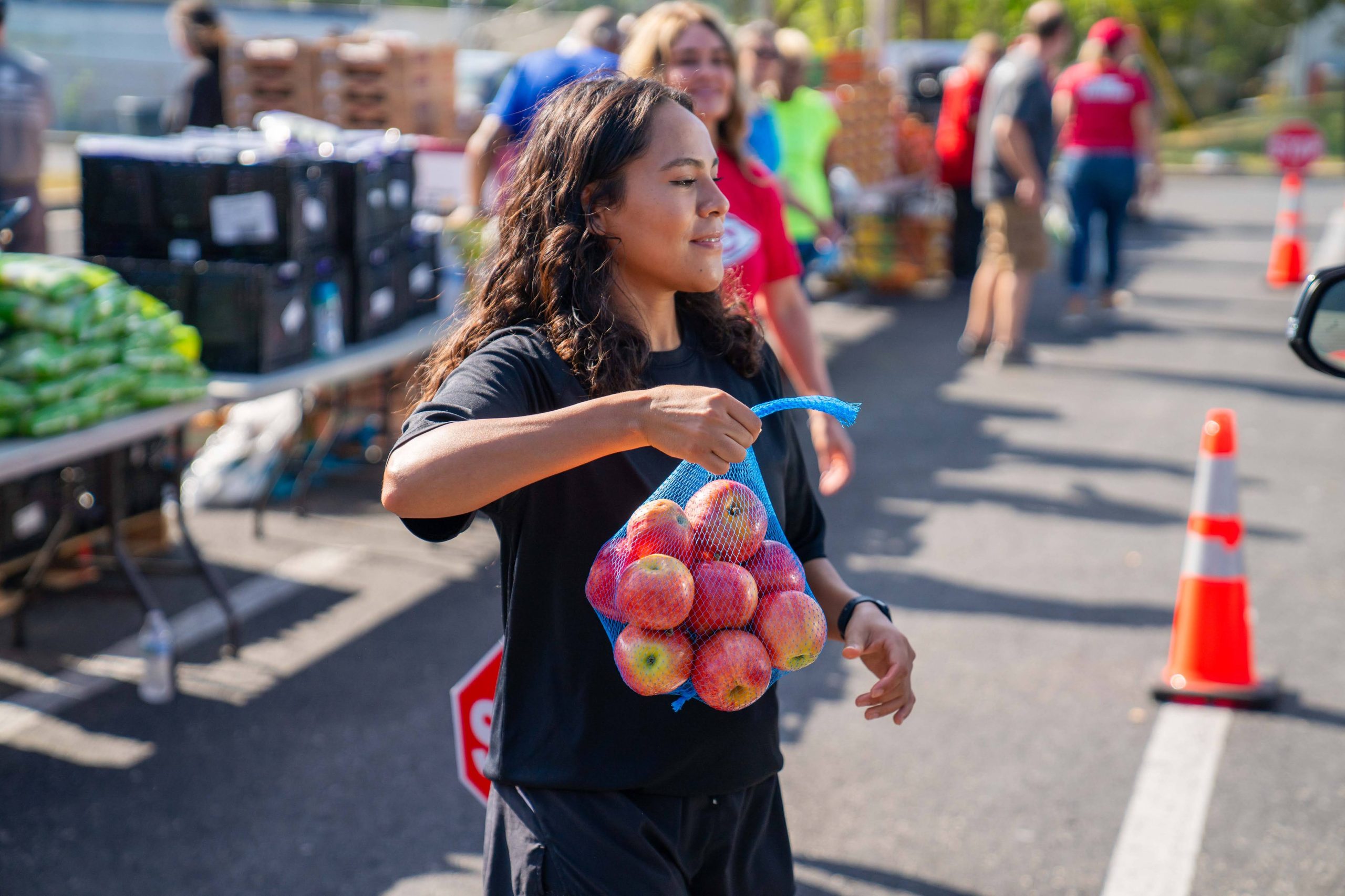 Smiling volunteer carrying a mesh bag of apples at an outdoor community food distribution event