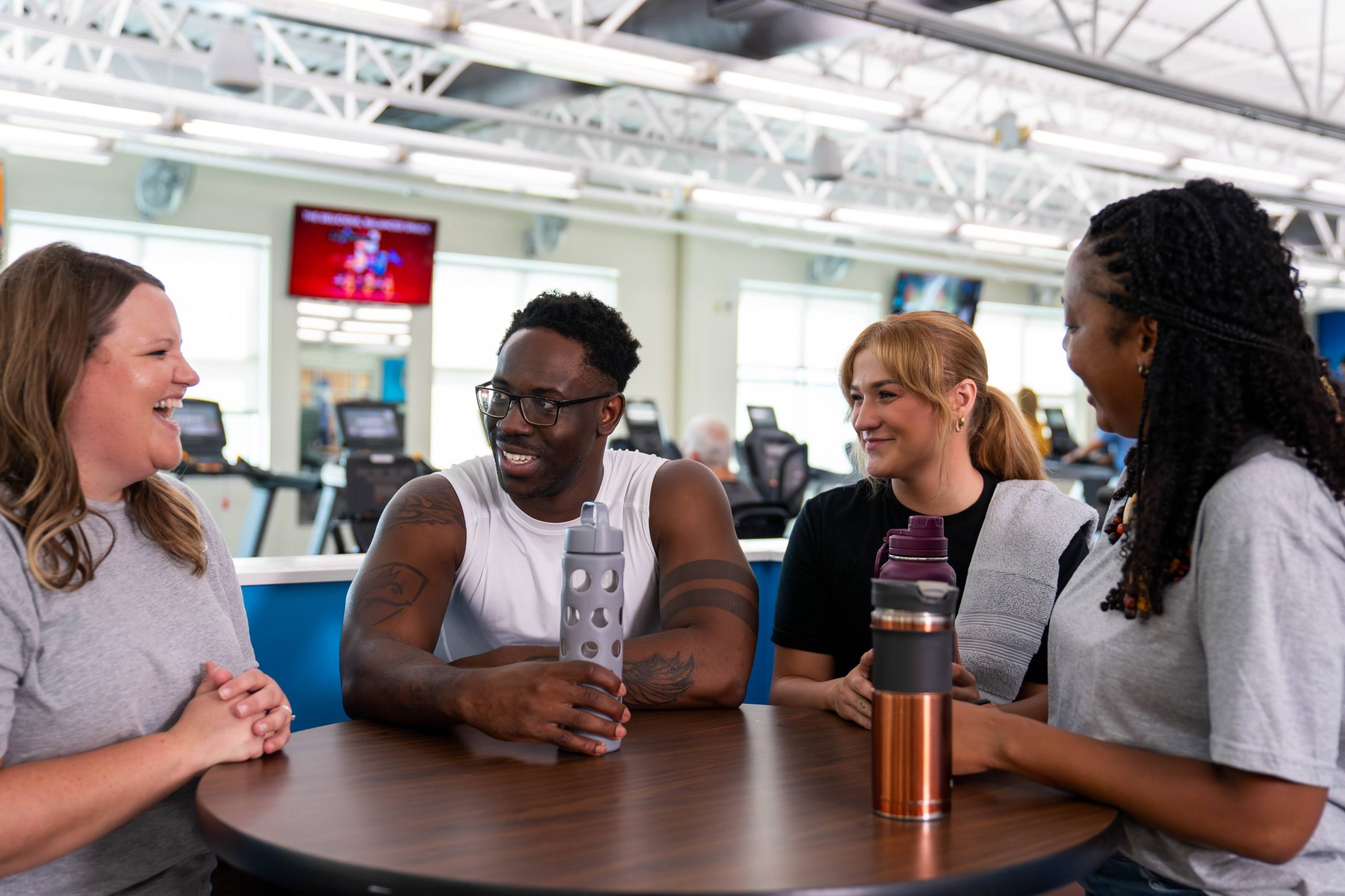 Four adults laugh together around a table with water bottles inside a bright fitness center
