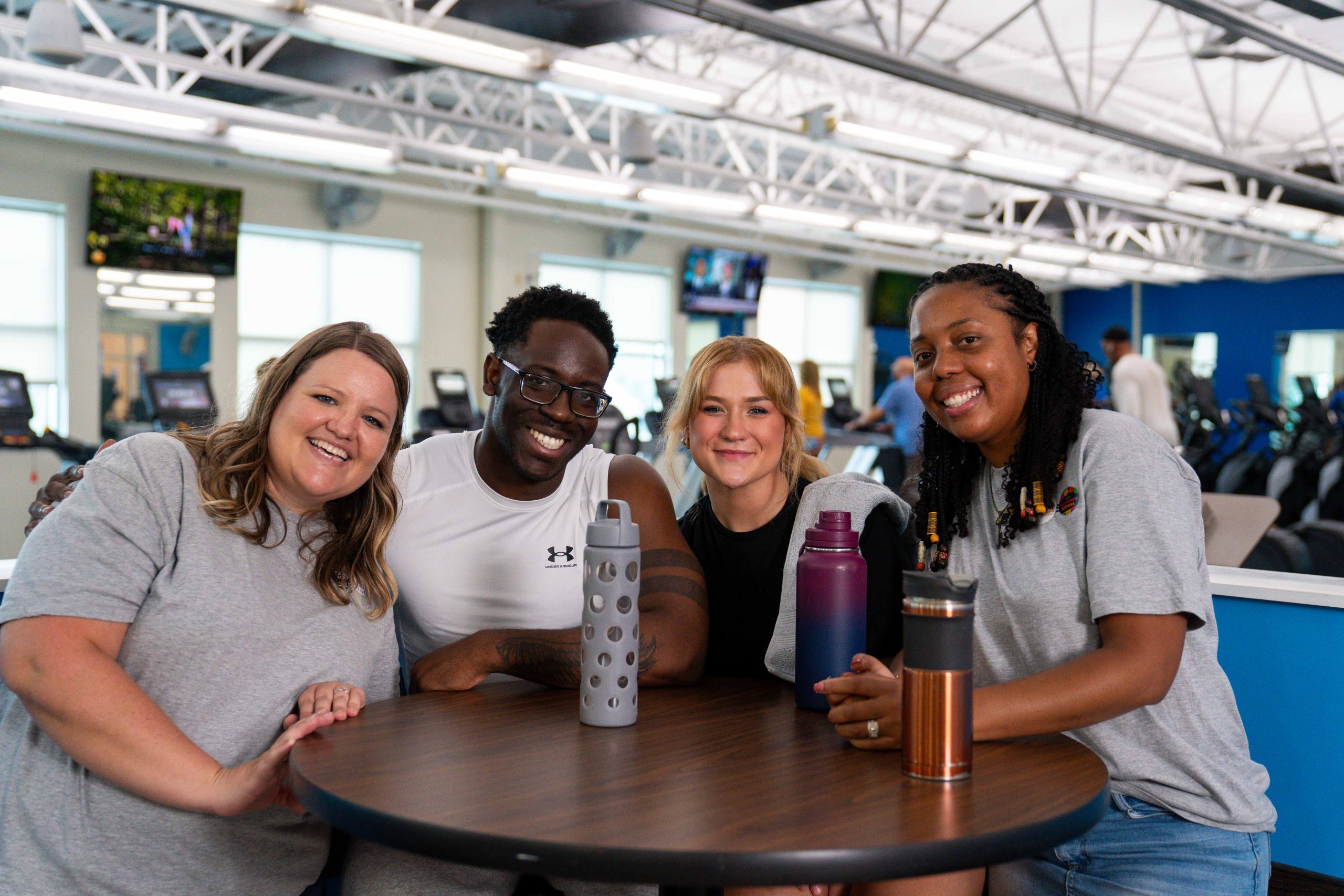 Four smiling adults with water bottles gather around a table inside a fitness center