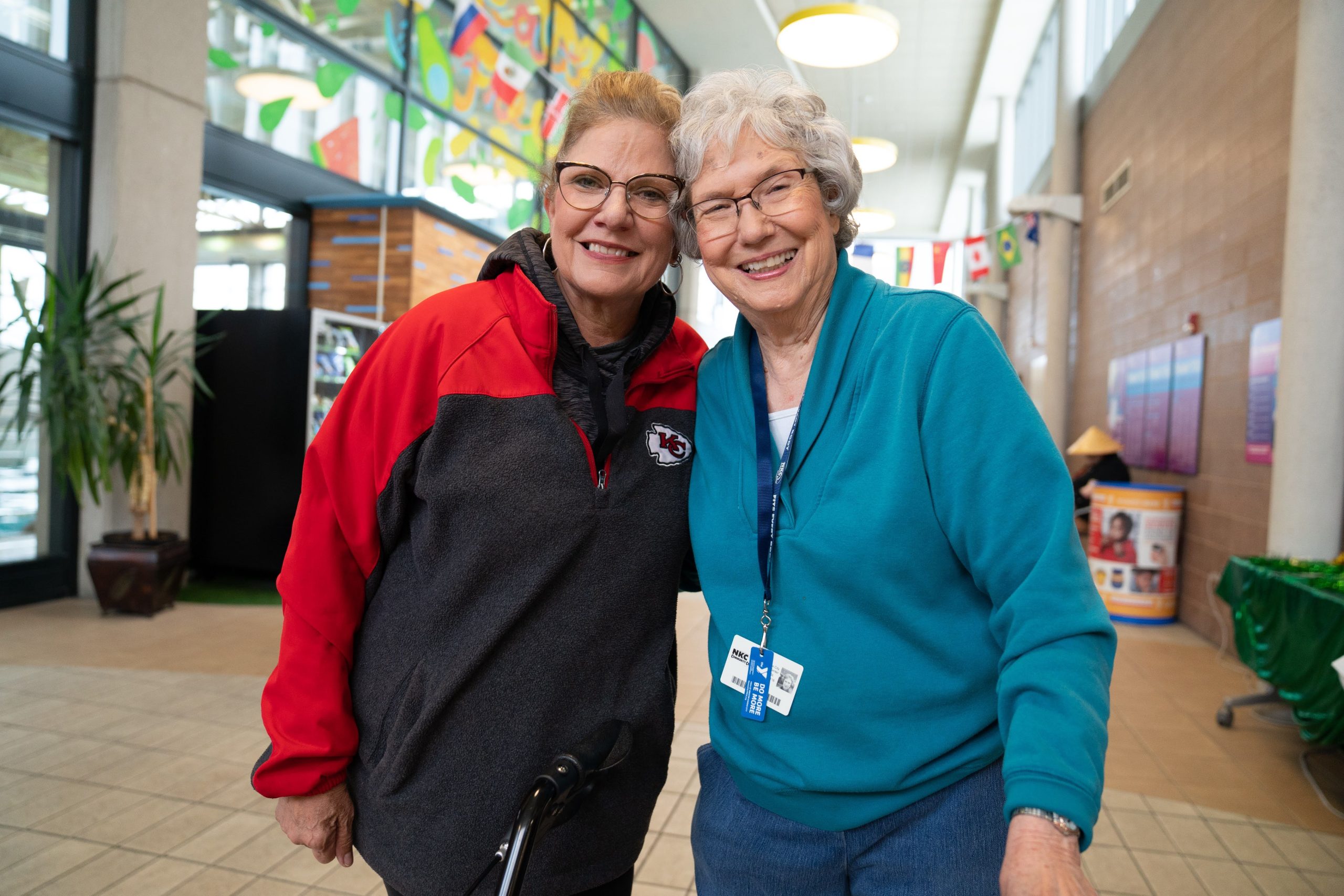 Two smiling older women stand arm in arm in a bright YMCA lobby, one wearing a staff lanyard