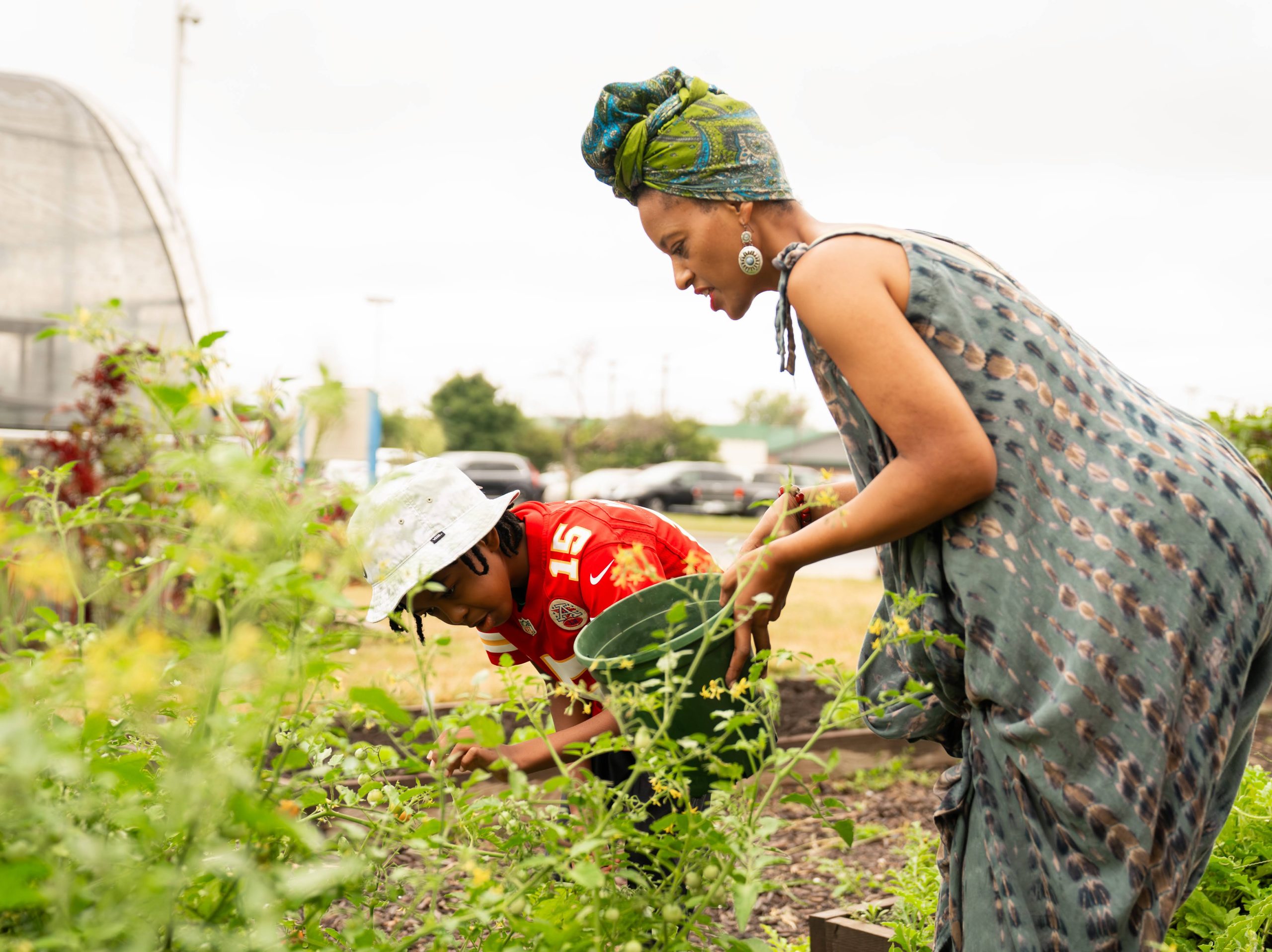 A woman in a headwrap and a child in a red jersey tend plants together in a community garden