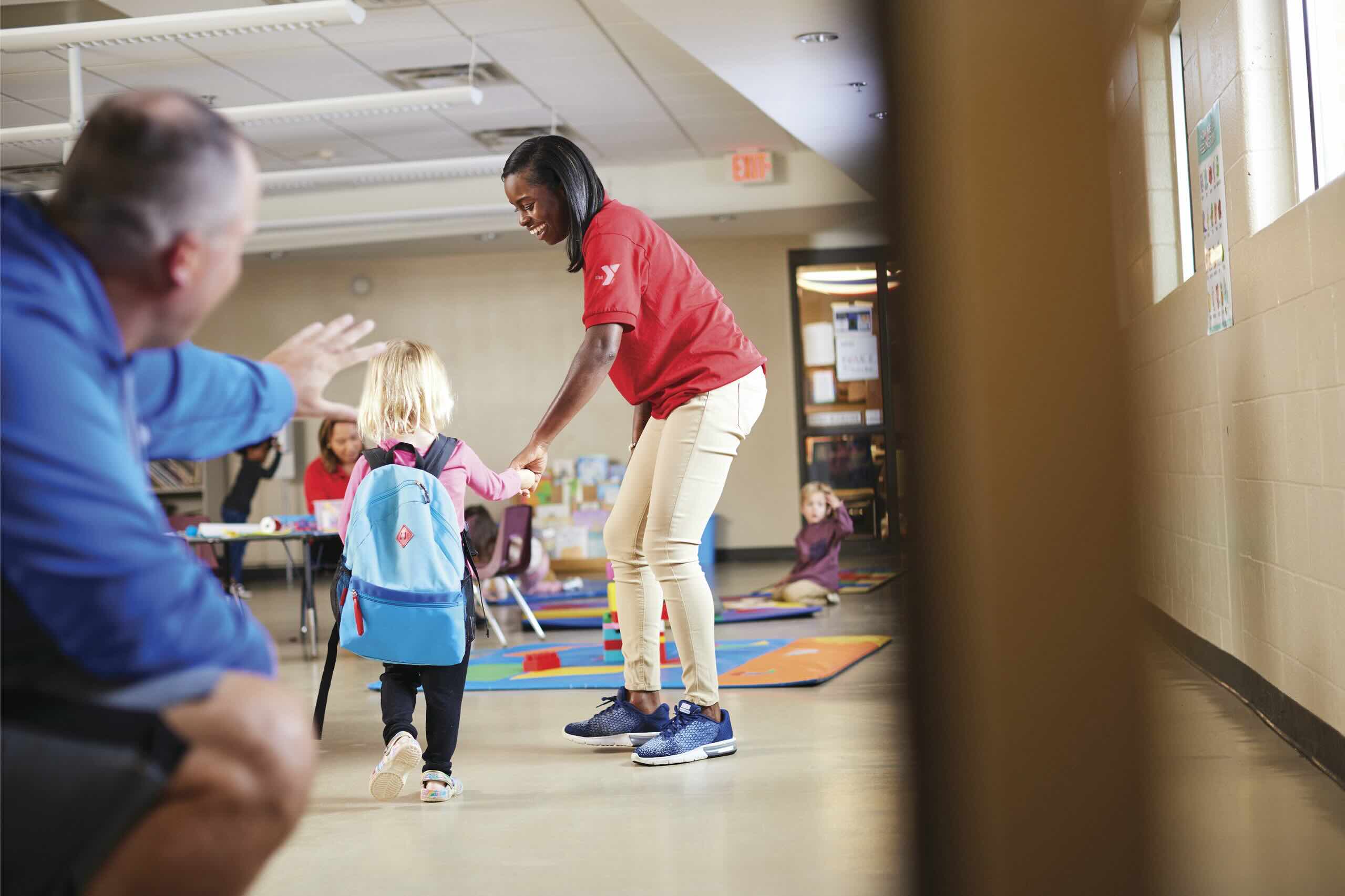 A YMCA staffer in a red polo walks a young child with a backpack toward a waving parent