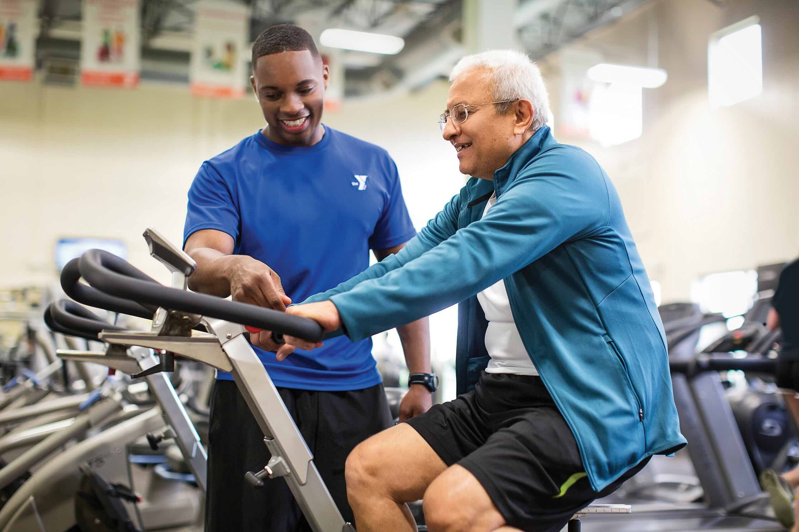 A young trainer in a blue Y shirt helps an older man adjust a stationary bike in a gym