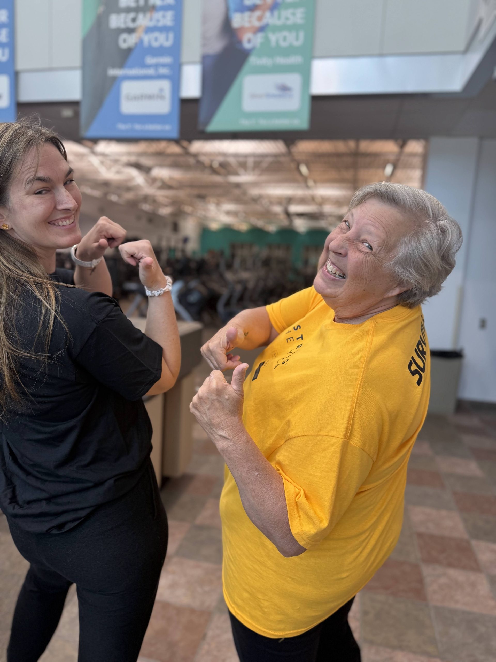 A younger woman and an older woman in a yellow shirt flex their biceps and laugh together