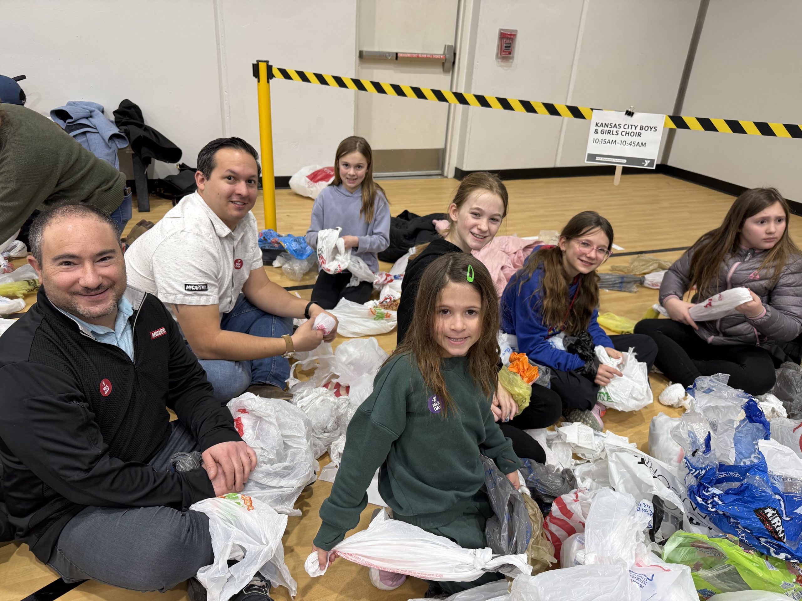 Adults and children kneel on a gym floor packing bags together during a service project