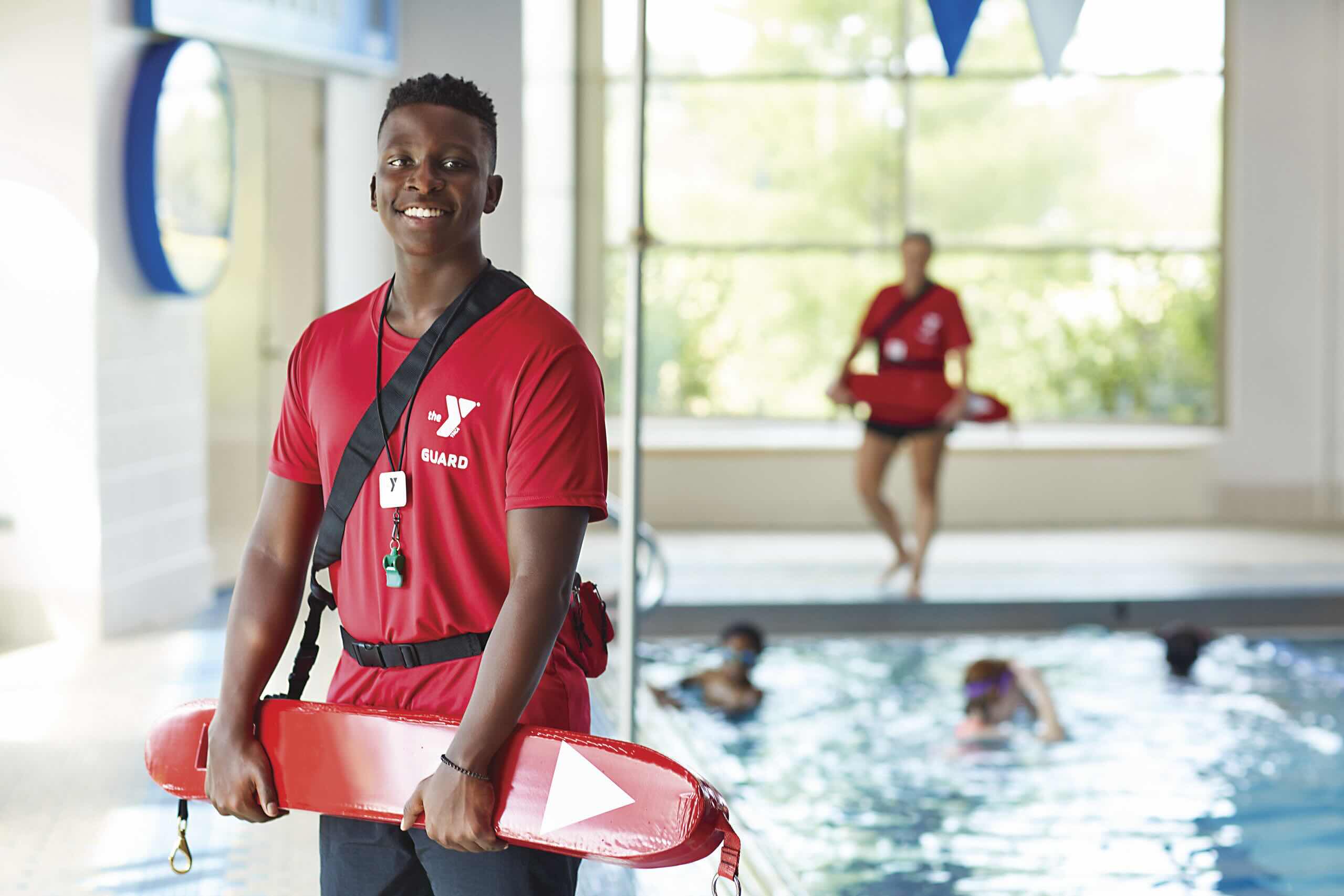 Smiling YMCA lifeguard in red guard shirt holding a rescue tube by an indoor pool