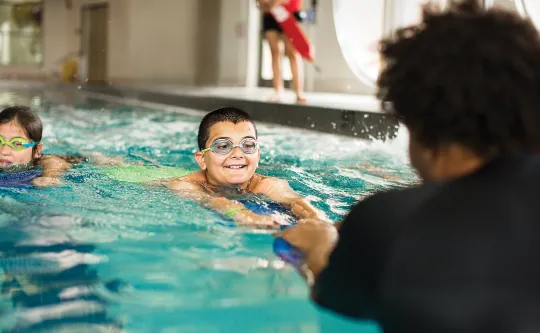 Smiling boy in goggles swimming toward an instructor during a pool swim lesson