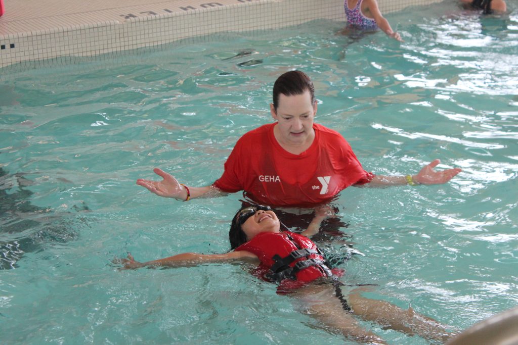 YMCA instructor in red shirt supports a child floating on their back in the pool