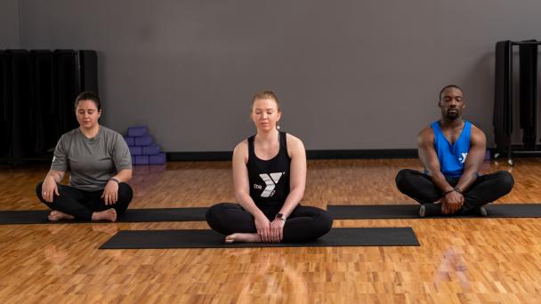 Three adults meditate cross-legged on yoga mats in a calm studio