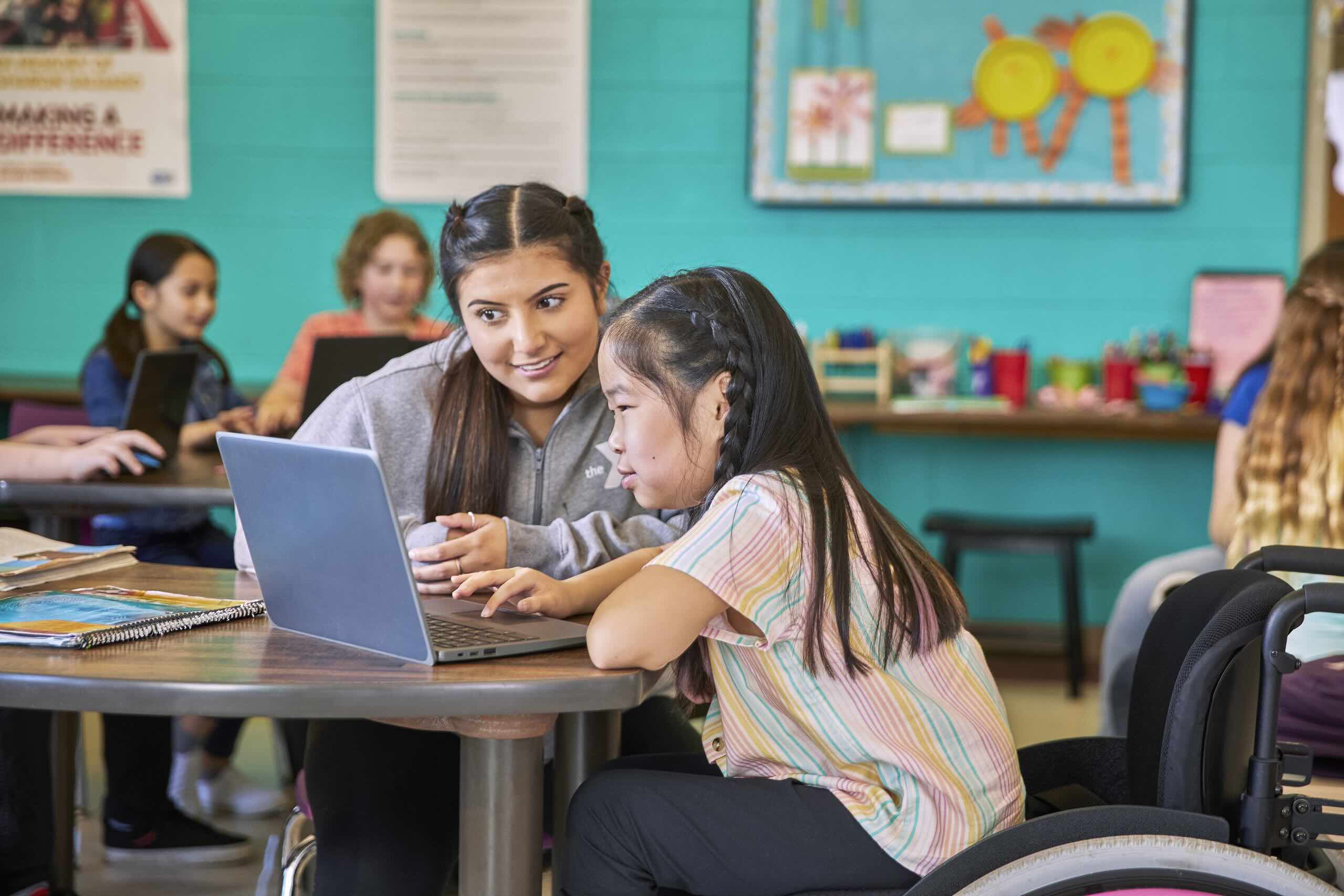 Teen mentor helping a younger student on a laptop in a colorful classroom