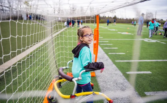 Smiling girl in glasses using a walker beside a soccer net on a turf field
