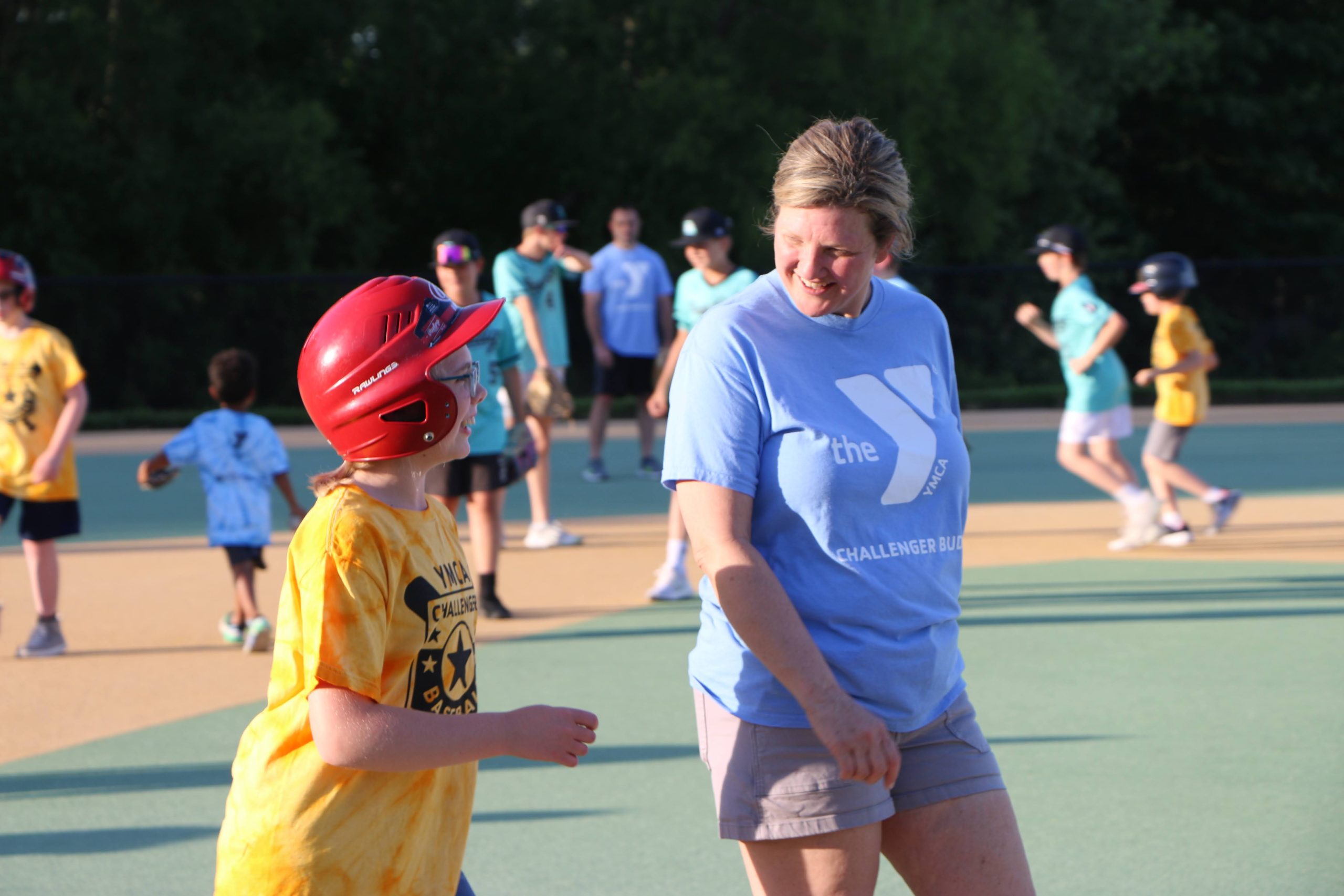 Adult volunteer in YMCA Challenger Buddy shirt smiling with a young ballplayer in a red helmet