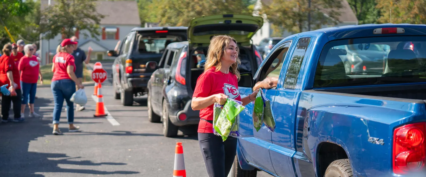 Smiling volunteer in red Chiefs shirt loading grocery bags into vehicles at a food drive