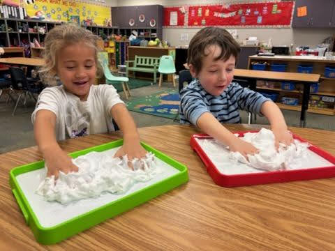 Two preschoolers laughing while playing with shaving cream on trays in a classroom