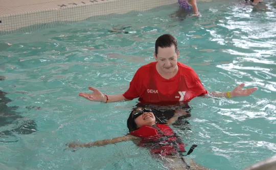 YMCA instructor in red shirt supporting a smiling child floating on their back in a pool