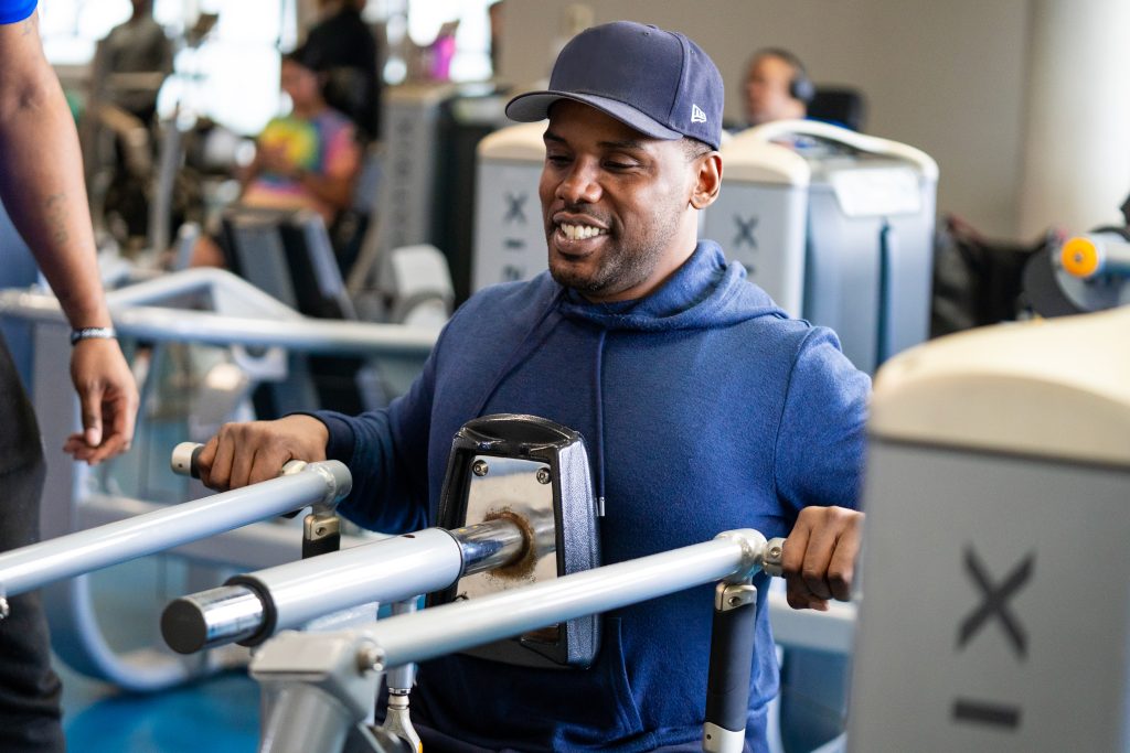 Smiling man in a navy hoodie and cap using a chest press machine on a busy gym floor