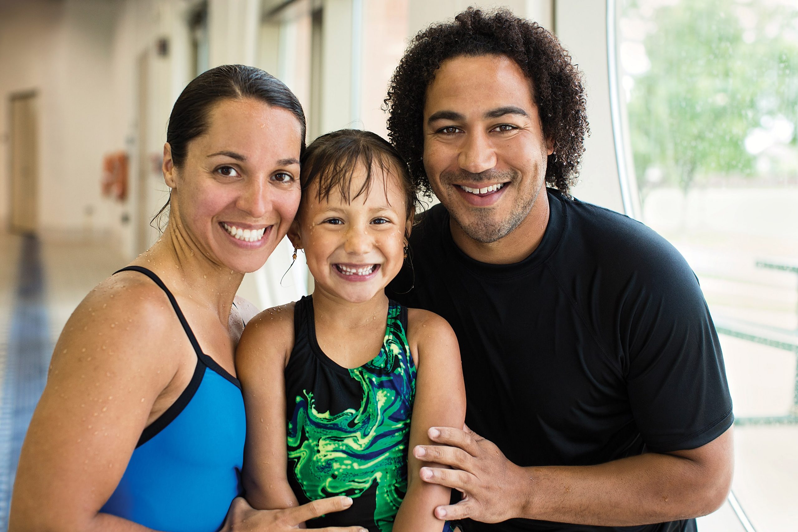 Smiling mother, young daughter, and father in swimwear together at an indoor pool
