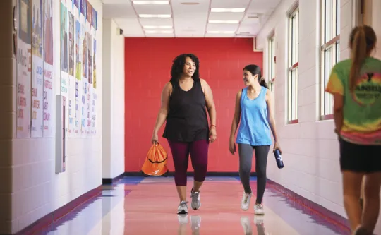 Two women in workout clothes walking and chatting down a bright YMCA hallway with a red accent wall