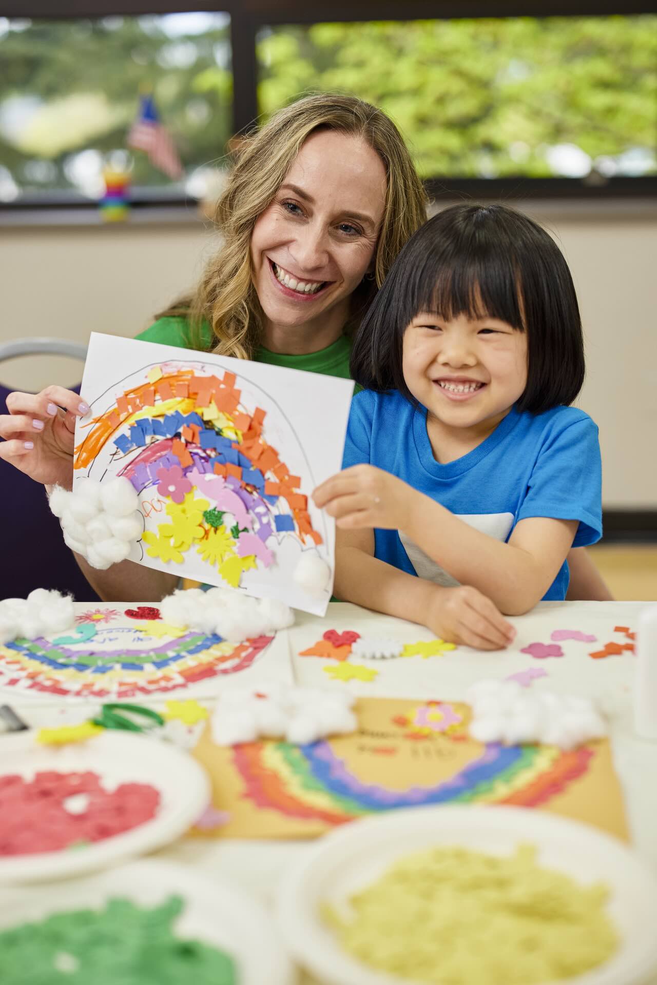 Smiling woman and young child holding up a colorful rainbow craft at a table