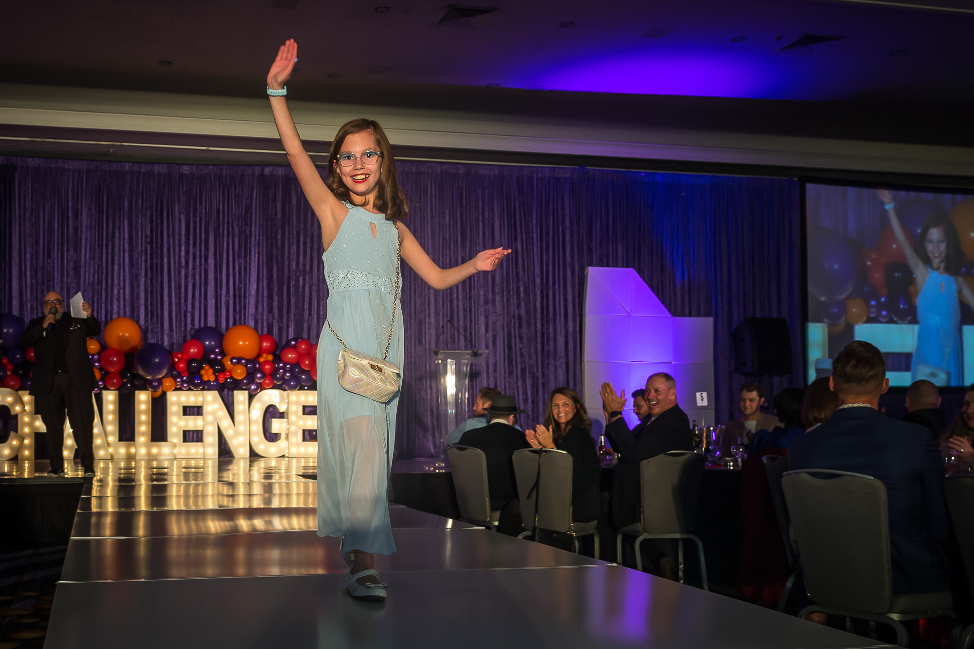 Challenger athlete Maddie Richardson struts down the runway in a floor length blue gown and a cute white purse. The audience is smiling and clapping.