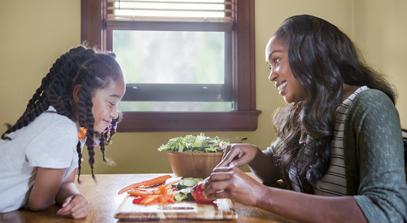 Mother and daughter smile while sitting at a table cutting up ingredients to put in a salad.