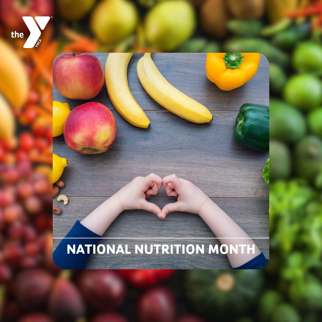 Child holding their hands in the shape of a heart on a table covered in a rainbow of fruit and vegetables. Text: "National Nutrition Month".