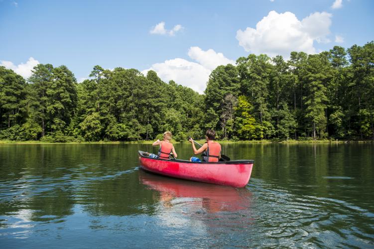 Two kids in lifejackets paddle a red canoe across a lake toward a tree-covered shoreline.