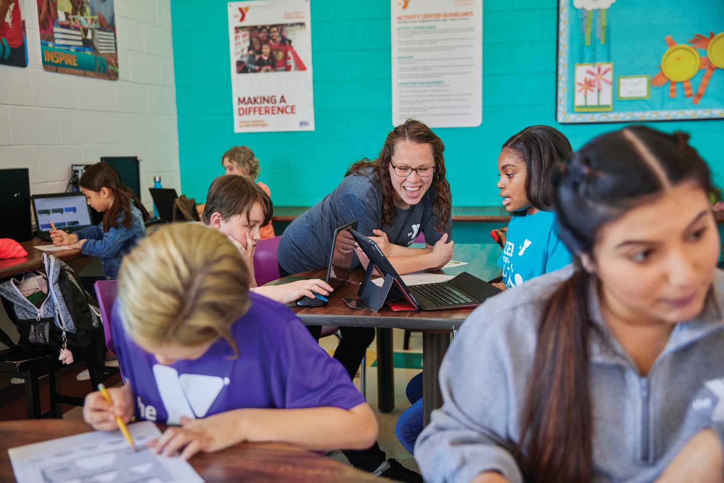 A Y staff member smiles while helping a young girl on her laptop computer in a classroom full of kids.