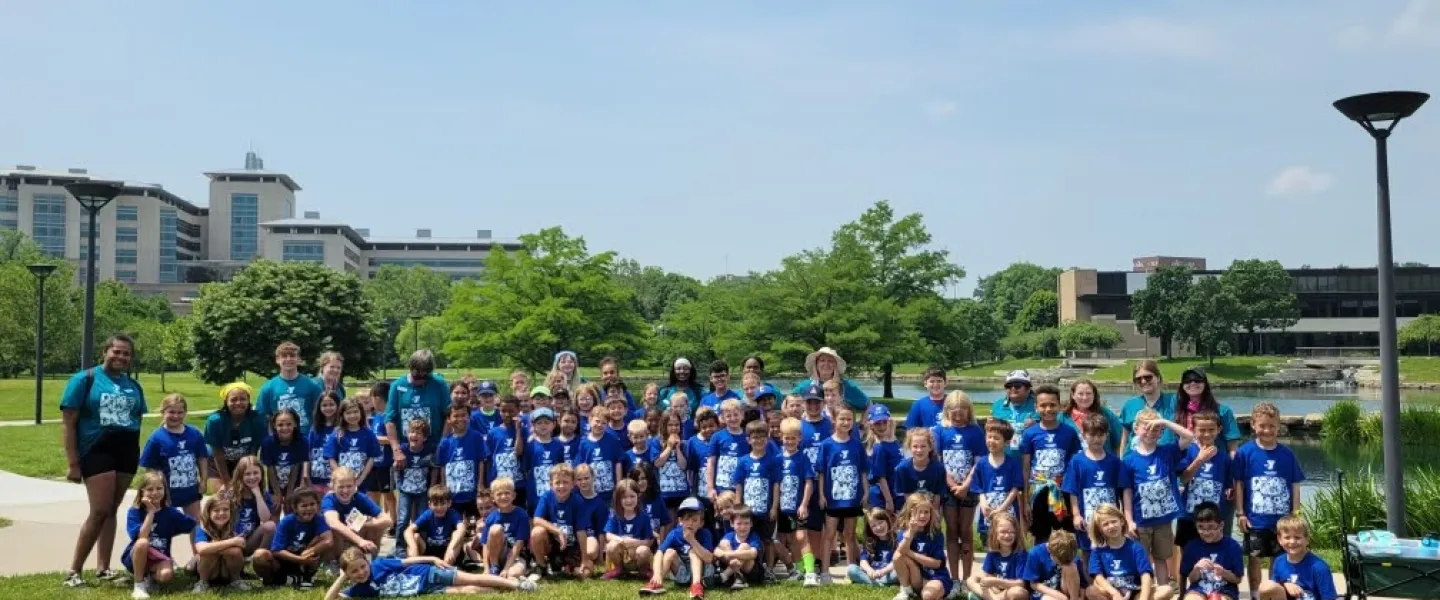 Large group of summer campers in blue shirts and counselors posing outdoors by a lake