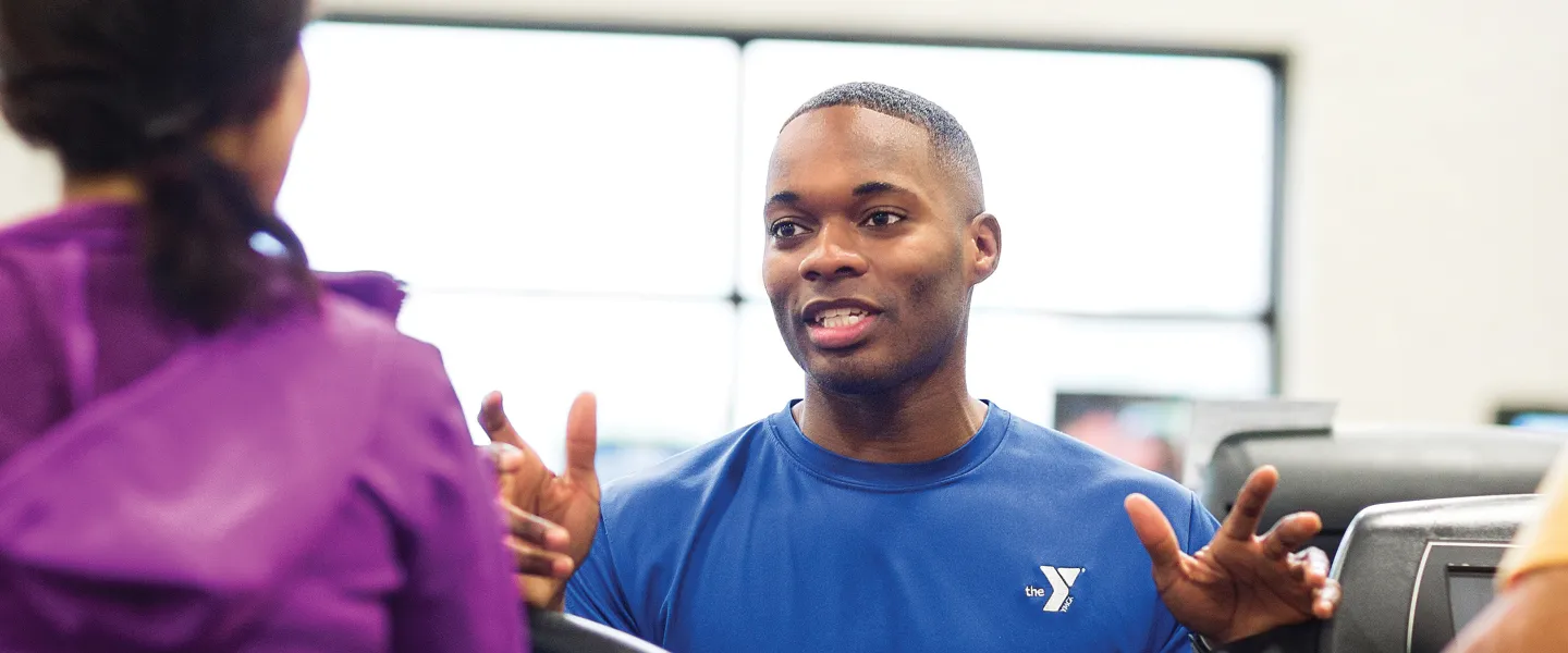 YMCA personal trainer in blue shirt gesturing and talking with members near cardio equipment