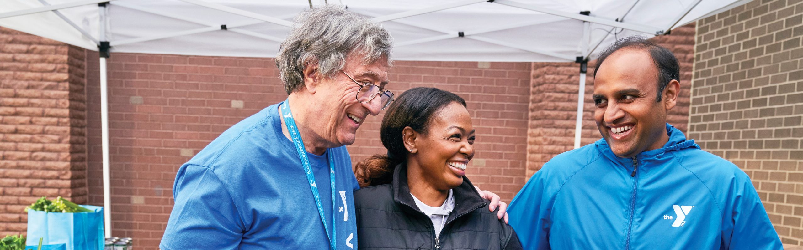 Three YMCA volunteers in blue jackets laughing together under a tent at an outreach event