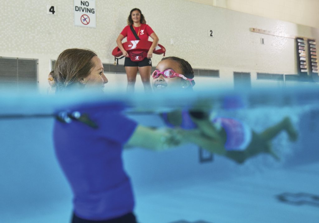 Instructor guides a young child in pink goggles swimming in an indoor pool