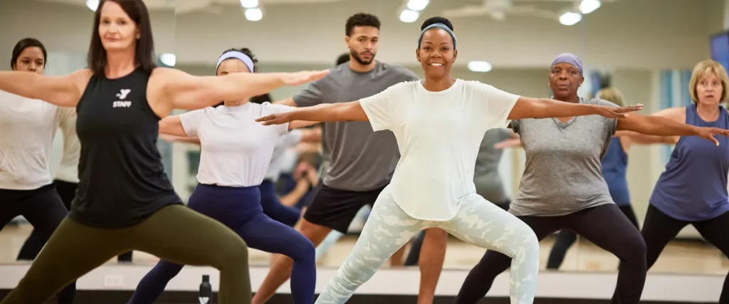 Diverse adults hold a yoga warrior pose in a bright fitness class led by staff