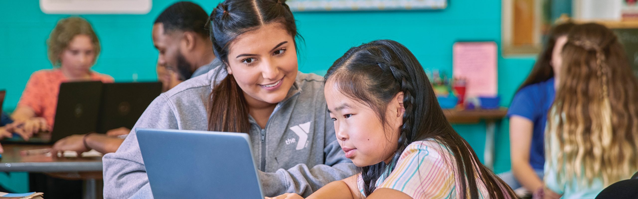 YMCA staff member helping a young girl on a laptop in a colorful classroom
