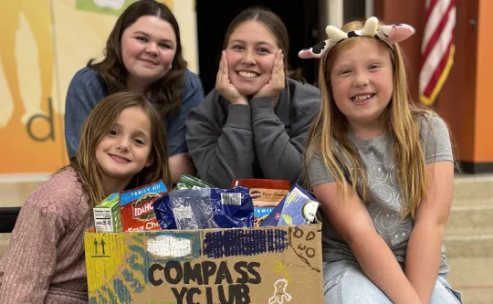 Four smiling kids posing behind a decorated Compass Y Club box filled with snacks