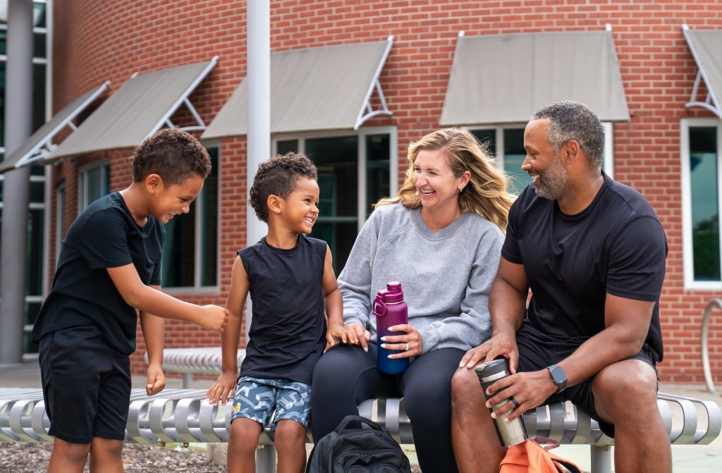 Family of four laughing together on an outdoor bench outside a brick YMCA building