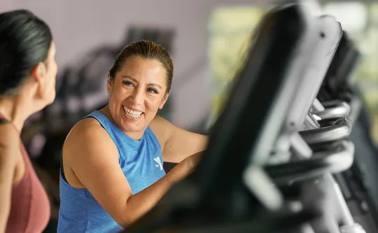 Smiling woman in blue tank top talking with another member beside gym cardio equipment