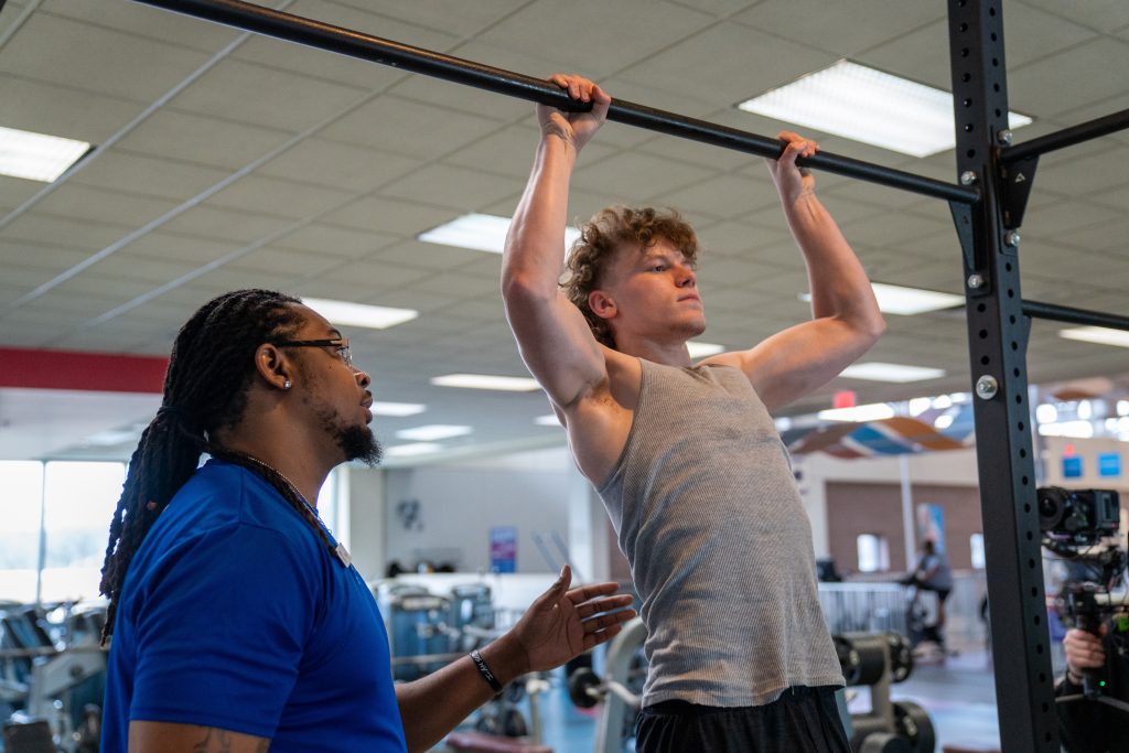 Trainer in blue shirt coaching a young man doing pull-ups on a gym rack