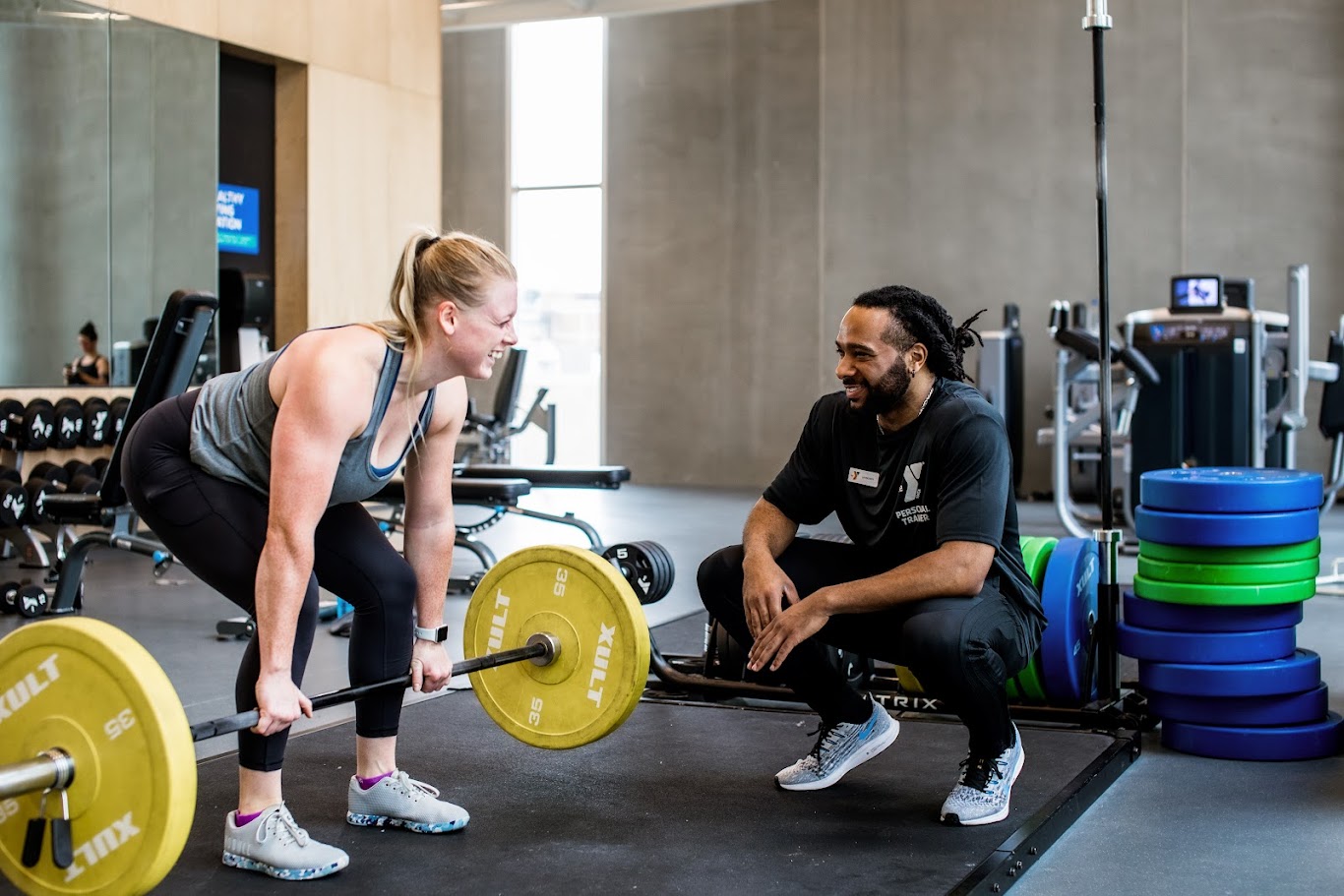 Smiling personal trainer coaches a woman lifting a barbell in a weight room