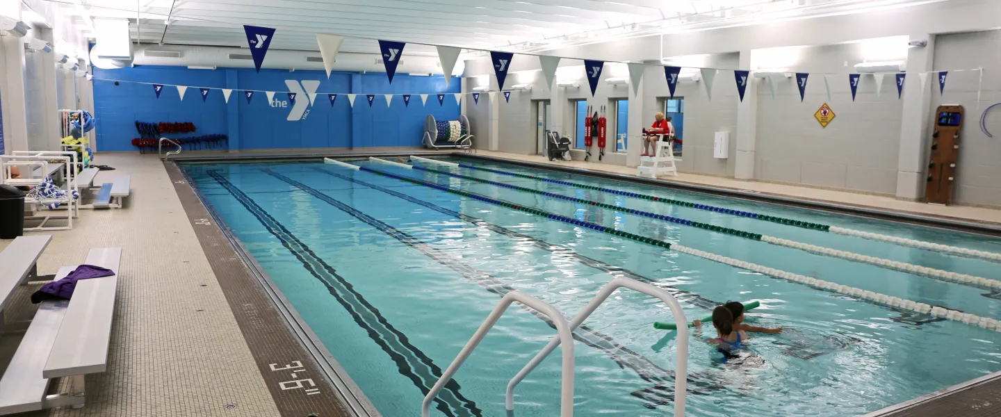 Indoor lap pool with YMCA banner flags, a lifeguard, and a child swimming with a noodle