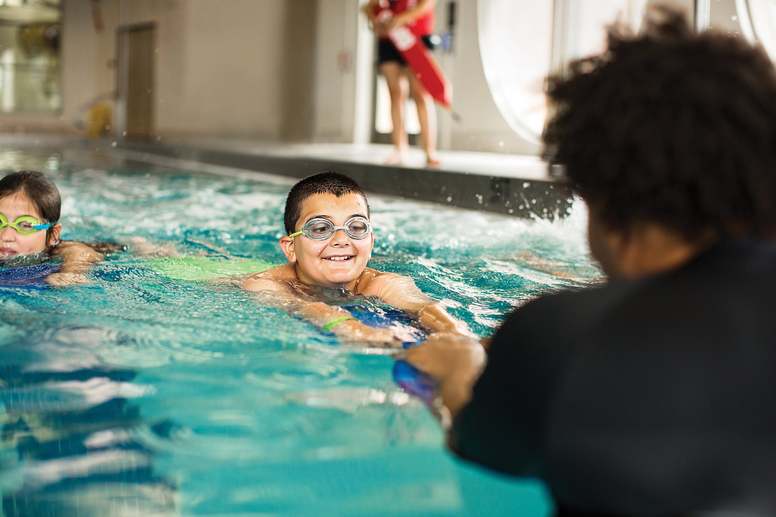Smiling boy in goggles during a swim lesson with an instructor at the pool edge
