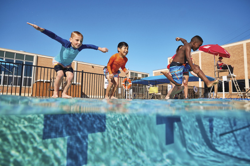 Three young boys jumping into an outdoor pool with a lifeguard watching